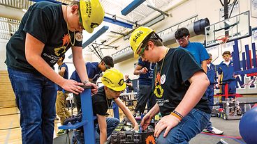 Teams of STEM students working on robotic projects inside a school gymnasium.