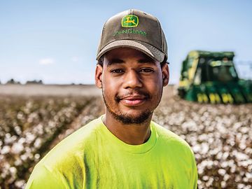 Farmer standing in cotton field with a John Deere harvester in the background.
